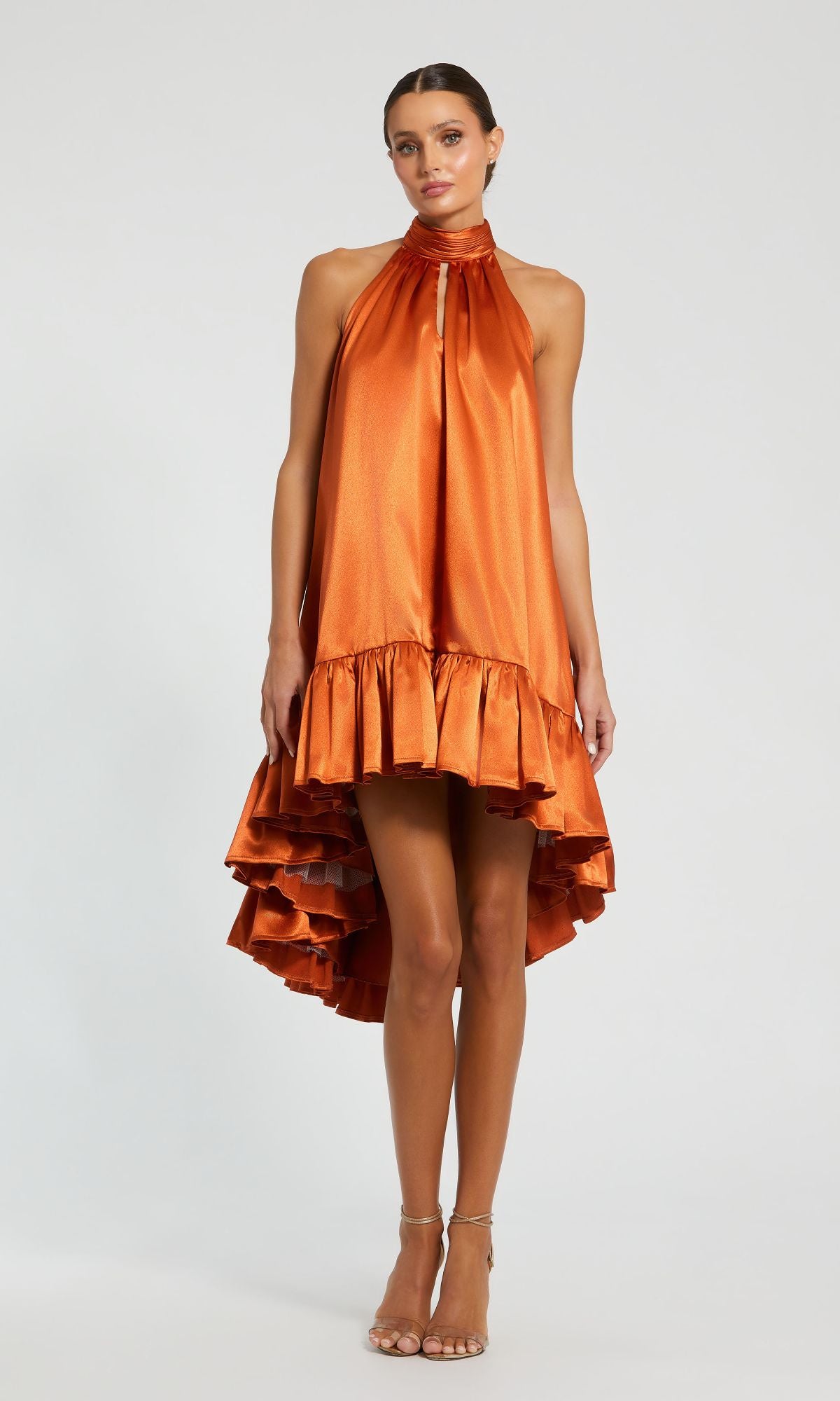 A woman stands in heels against a plain background wearing the Mac Duggal 20711 Satin Wedding Guest Dress—an orange, shiny, sleeveless piece with a halter neck and ruffled high-low hemline.